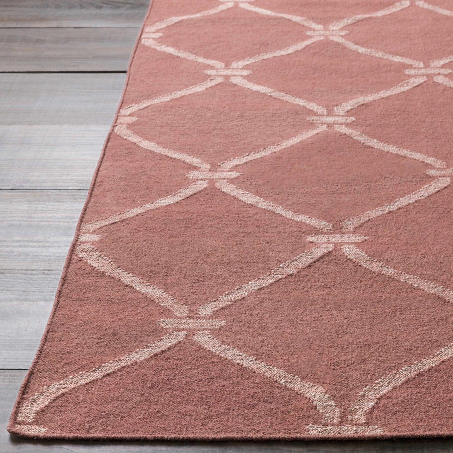 Close-up of a dusty pink Hazelhurst Area Carpet with a light gray diamond trellis pattern on a gray wood floor. Front