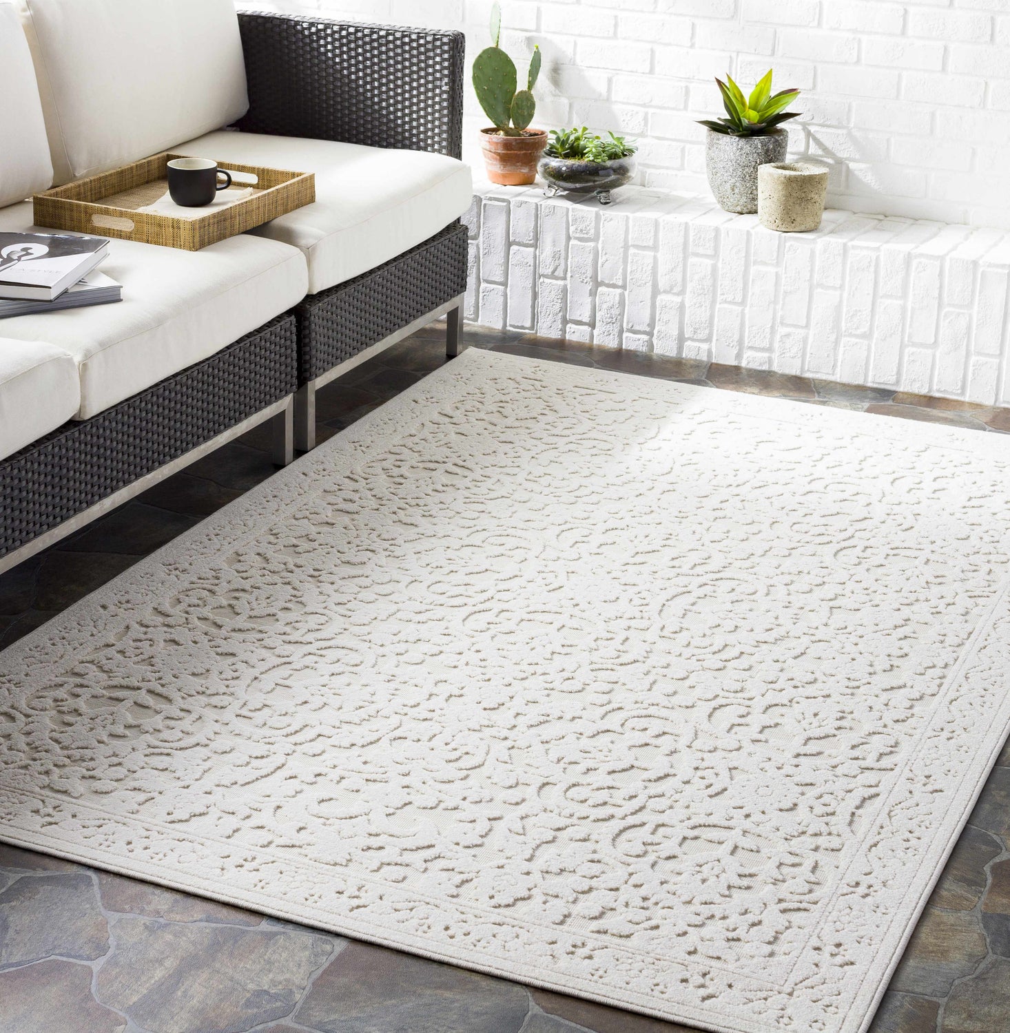 Bohemian patio with beige Innaloo 3D Outdoor Area Rug, dark wicker sofa, and white brick accent wall. Roomscene-Rectangle