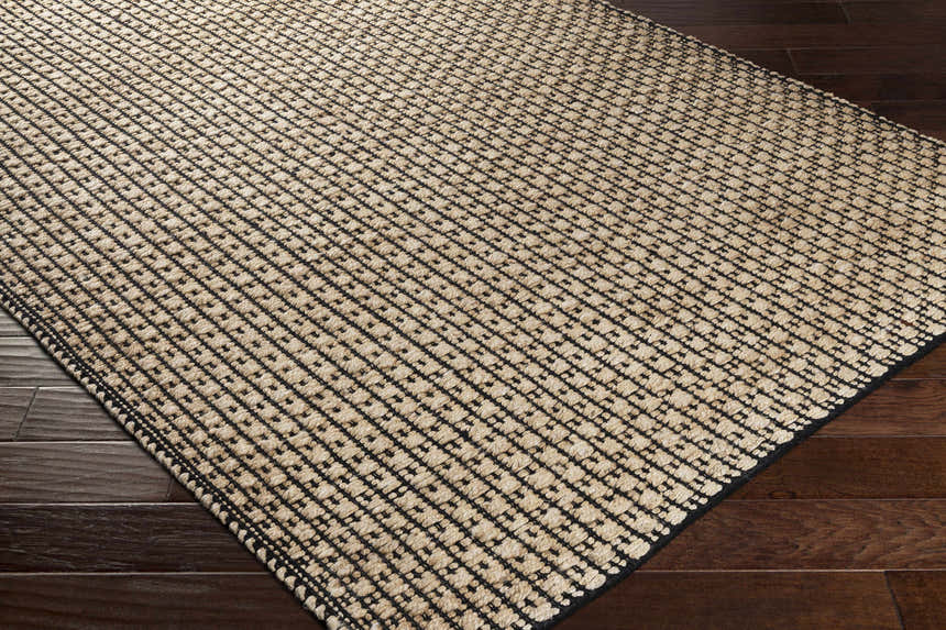 Close-up of Iqaluit Natural Jute Rug. Beige jute rug with a black grid pattern and border on dark wood floors. Corner