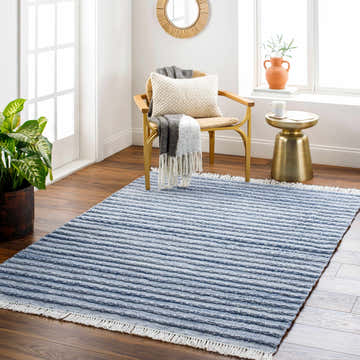 Light-filled room featuring the blue striped Jake Area Rug, a woven chair, and gold accent table. Roomscene-Rectangle