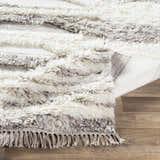Close-up of the hand-woven Kingsthorpe Area Rug. Cream and taupe shag rug with fringe detail on a wood floor. Fold