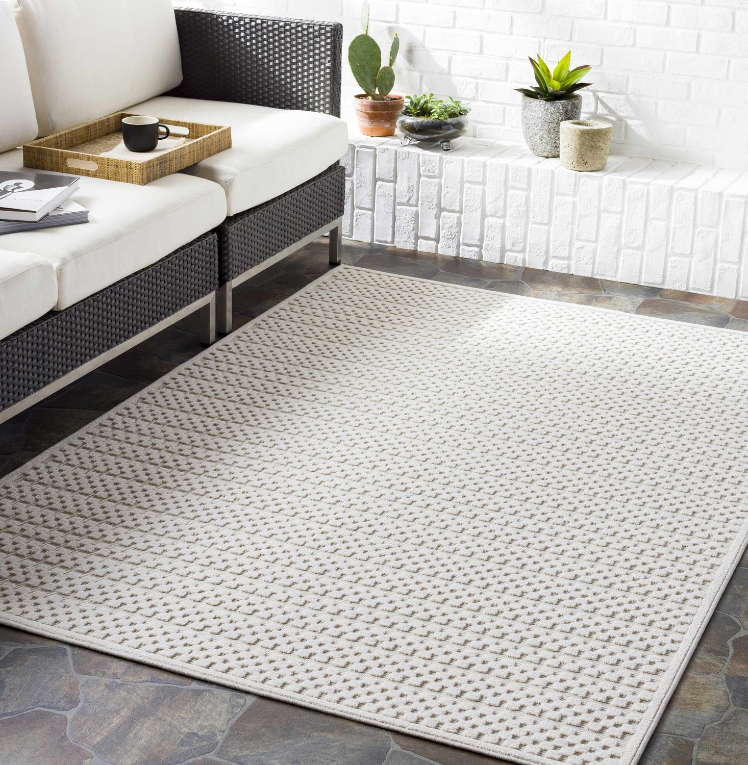Styled patio with a Kirkburton Beige Outdoor Rug, charcoal wicker sofa, and white brick wall. Roomscene-Rectangle