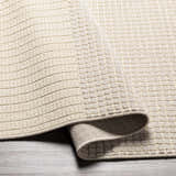 Close-up of the Lice Area Rug showing its textured, grid-like pattern and soft, neutral color on a gray wood floor. Fold