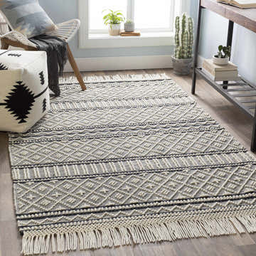 Living room scene with a black and white geometric Leon Area Rug, chair, pouf, desk, and plants. Roomscene-Rectangle