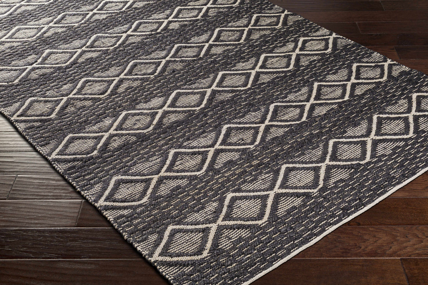 Close-up of the Maluanluan Jute Carpet. Hand-woven rug with black and light beige diamond pattern on dark wood floor. Corner