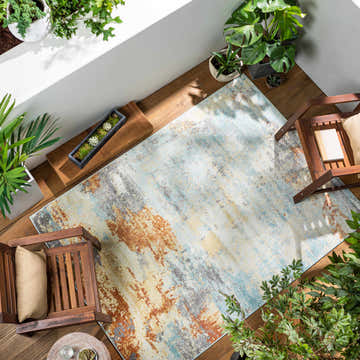 Sunlit patio with Mecosta Light Outdoor Area Rug. Wood floor, chairs, and potted plants create a relaxing coastal vibe. Roomscene-Rectangle