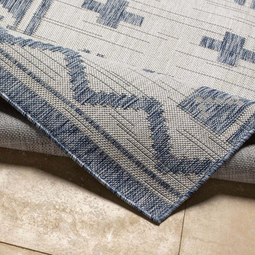 Close-up of the Mangoso Gray Area Rug showing its blue geometric pattern and woven texture on a beige tile floor. Fold