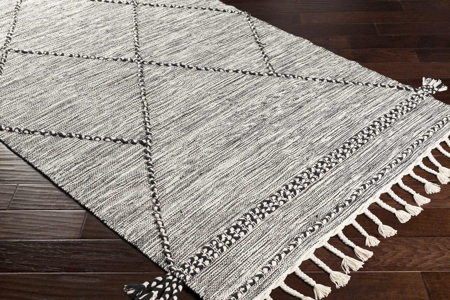 Close-up of Neal Black/Cream Trellis Cotton Rug with Tassels on dark wood floor. Hand woven rug with geometric pattern. Corner