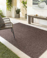 Outdoor patio scene with a Nuela Dark Brown Area Rug, wicker chair, bench, and potted plants. Roomscene-Rectangle