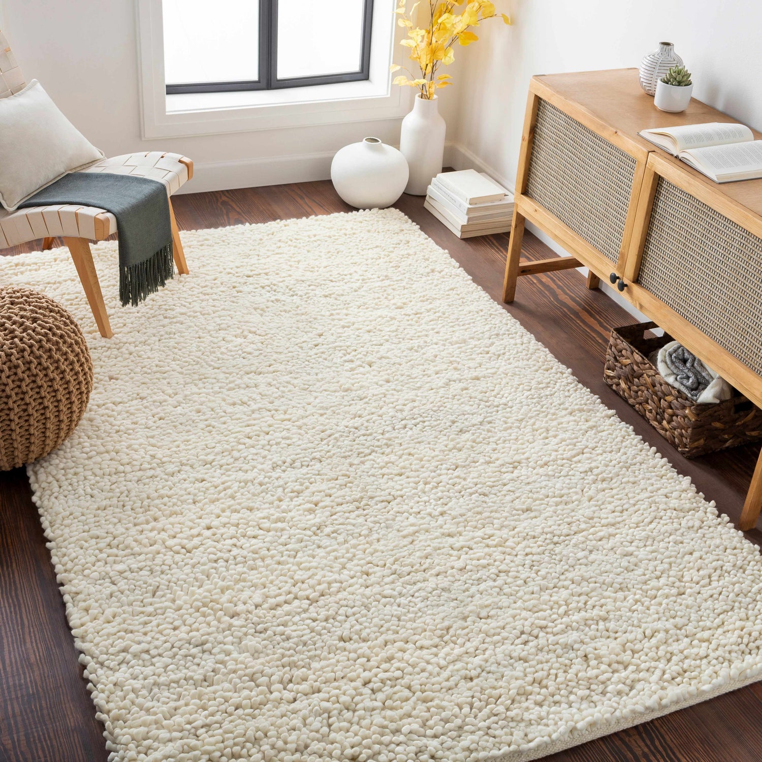 Living room scene with a cream Oakridge Area Rug, wood cabinet, chair, and woven pouf. Roomscene-Rectangle