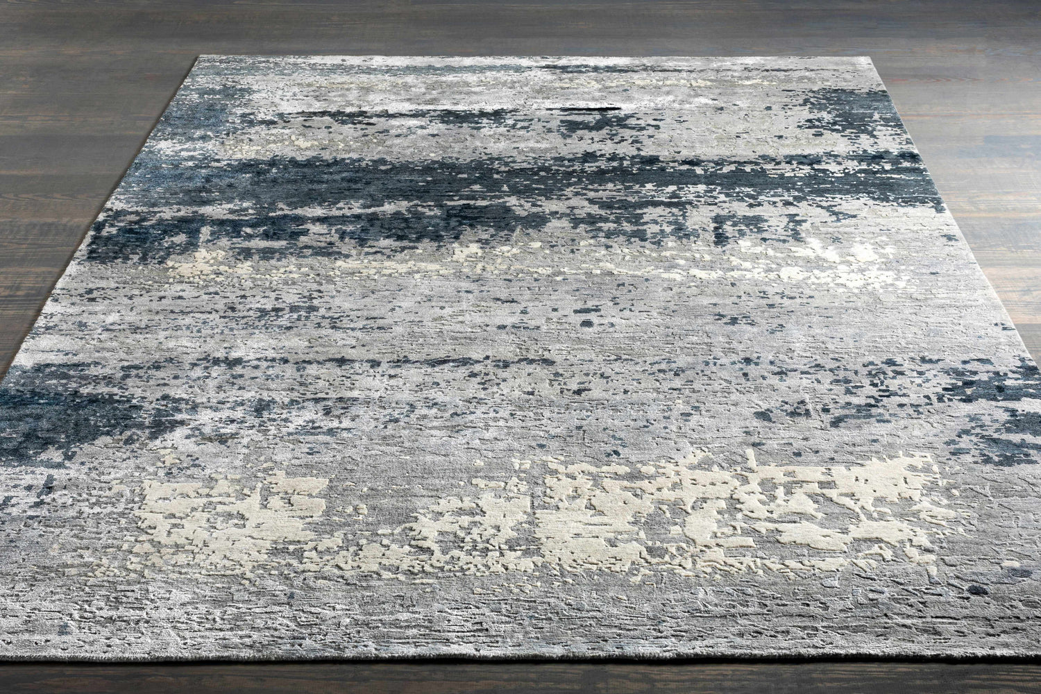 Close-up of the Okemos Area Rug. Contemporary design in charcoal, dark blue, gray, light beige, and teal on a wood floor. Styleshot