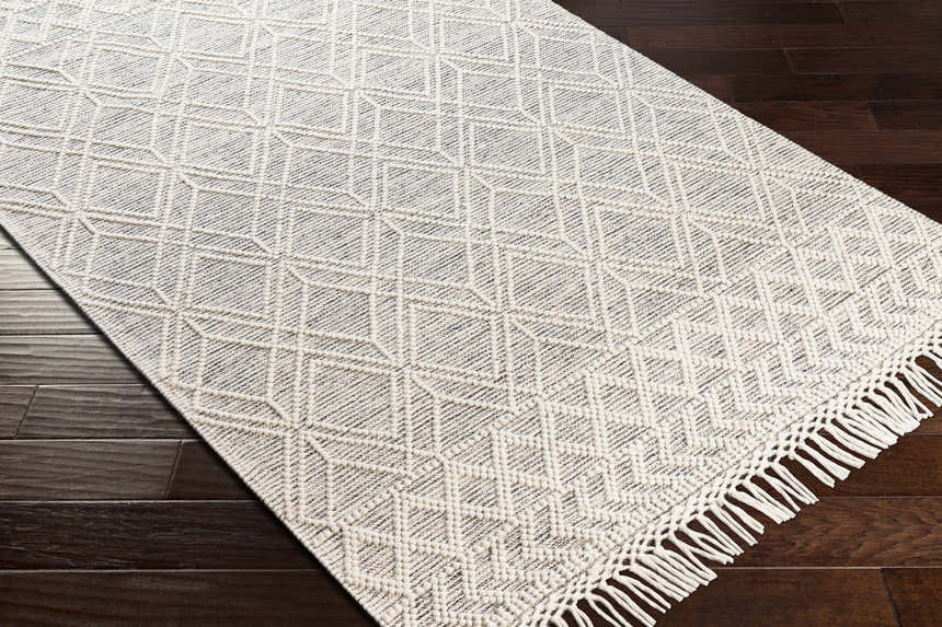 Close-up of the Ossun Wool Rug. Cream rug with geometric pattern and tassels on dark wood floor. Corner