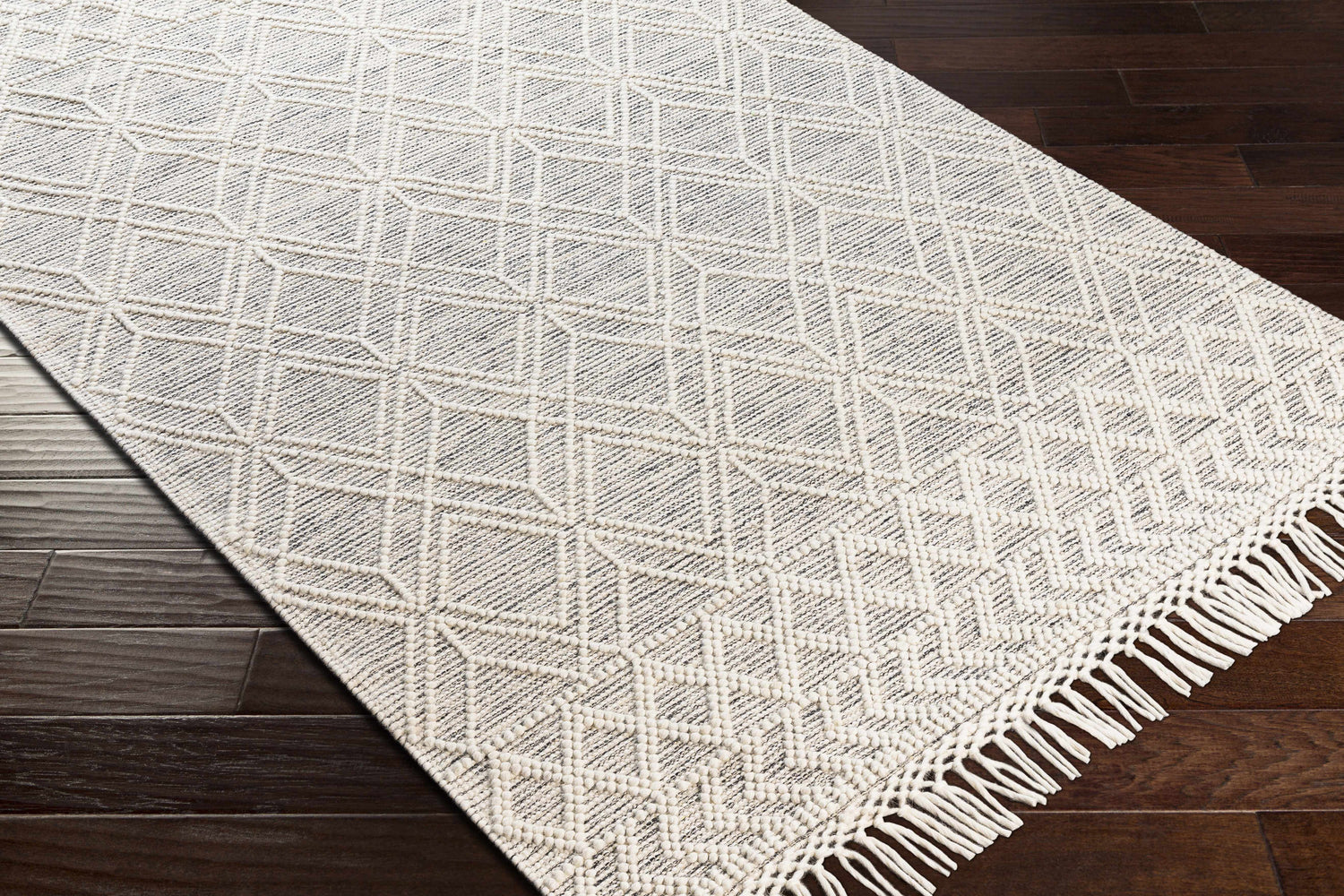Close-up of the Ossun Wool Rug. Cream rug with geometric pattern and tassels on dark wood floor. Corner