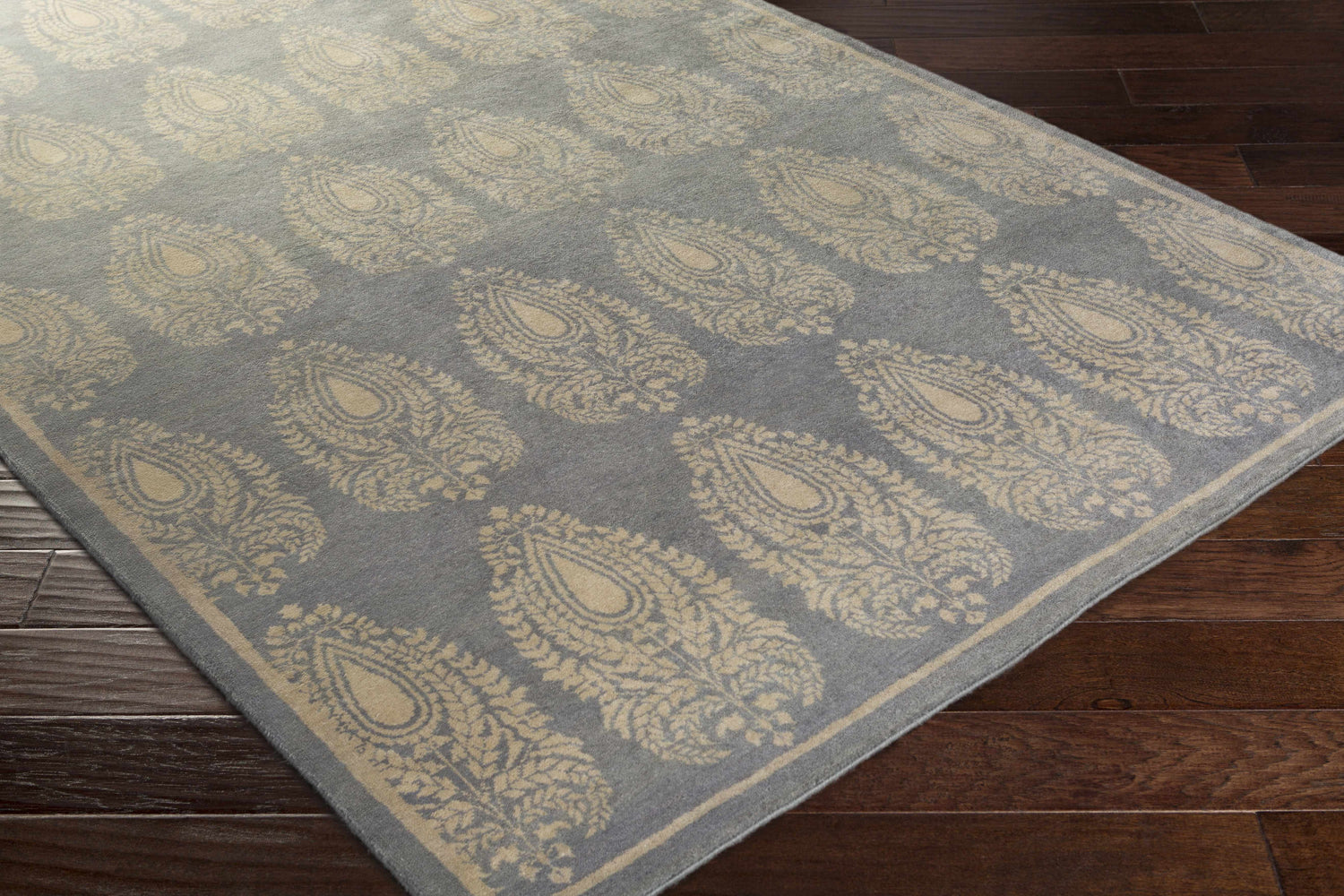 Close-up of the Otho Area Carpet. Gray wool rug with a taupe paisley pattern on a dark wood floor. Corner