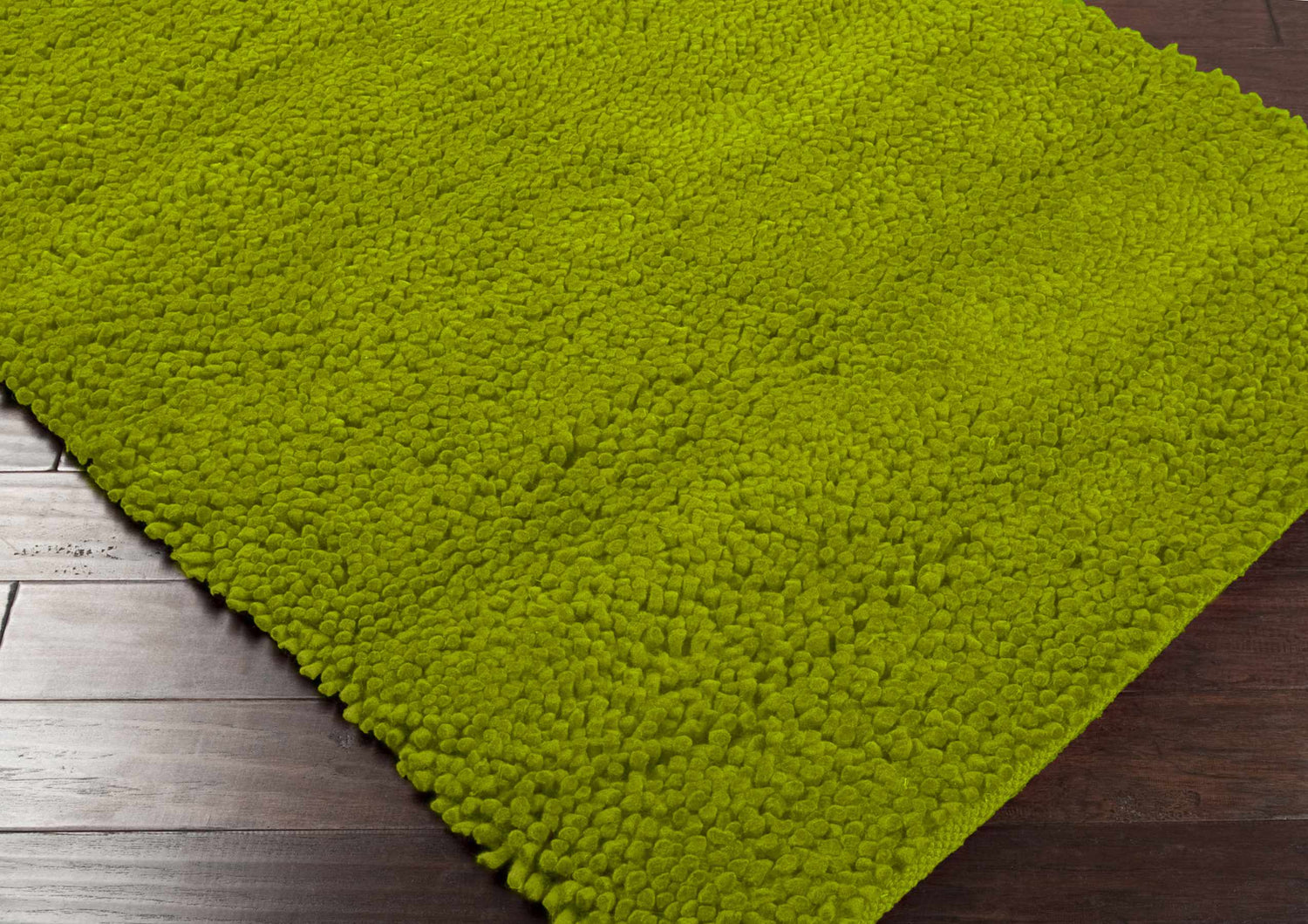 Close-up of a Frederika Solid Olive Green Wool Shag Rug showing its texture and color on dark wood flooring. Corner