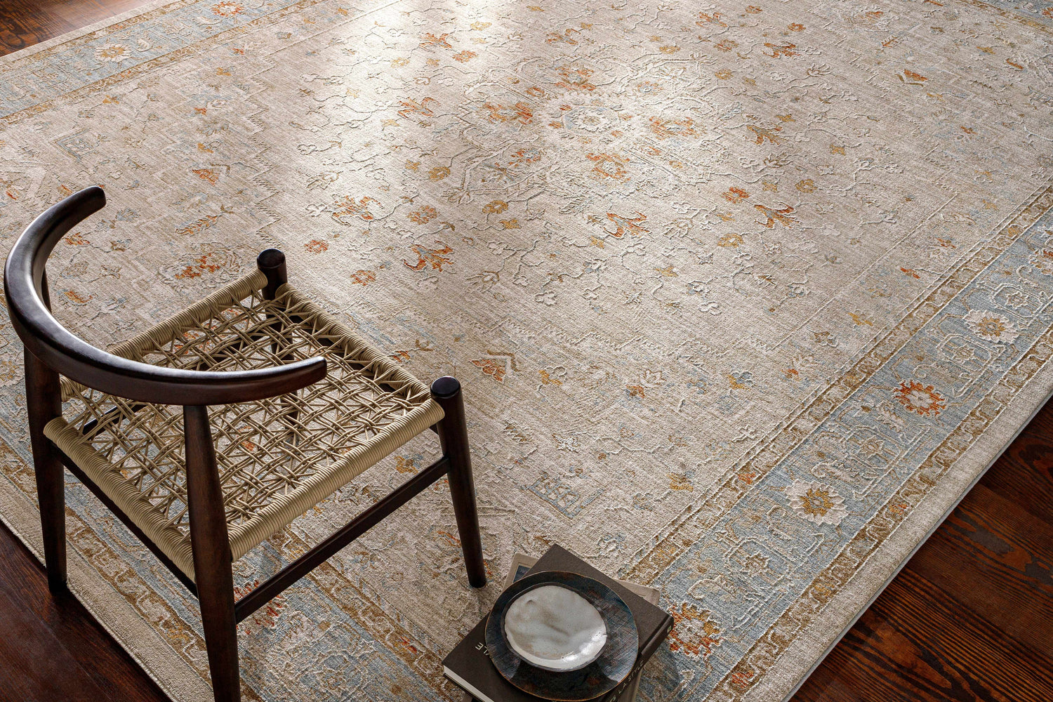 Room scene with Phyllis Thick Luxe Statement Rug in light beige, brick red, and denim colors. Woven chair and dark wood floor. Styleshot