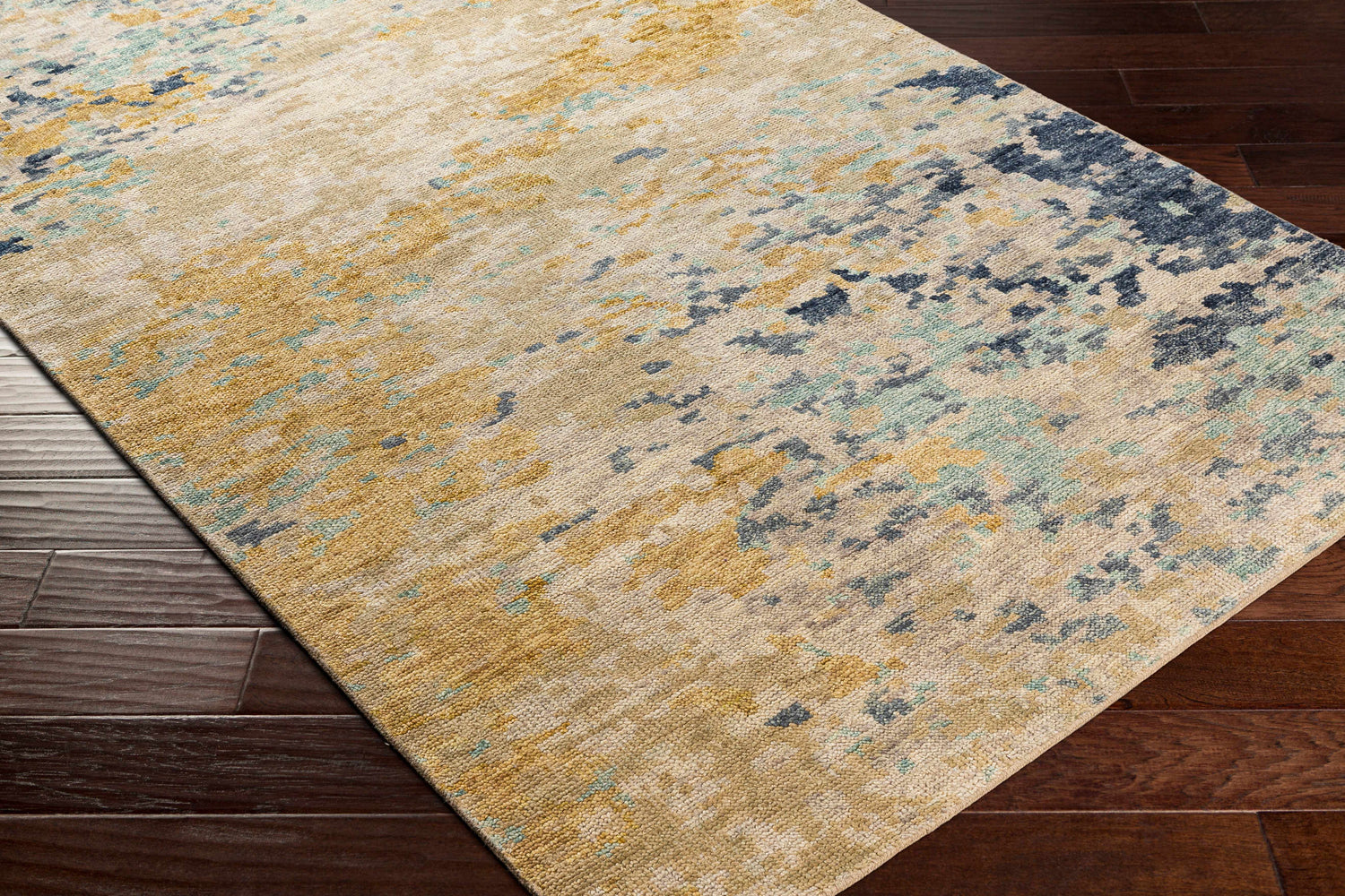 Close-up of the Pias Area Rug showing its beige, gray, and blue contemporary pattern and hand-knotted texture on dark wood floor. Corner