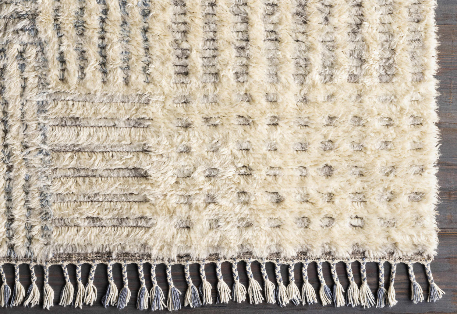 Close-up of the hand-knotted Plean Premium Rug. Cream wool rug with gray geometric patterns and braided tassels. Corner