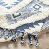 Close-up of the Painesdale Blue/White Wool Carpet. Hand-woven rug with geometric pattern, fringe, and blue/white braided tassels. Fold