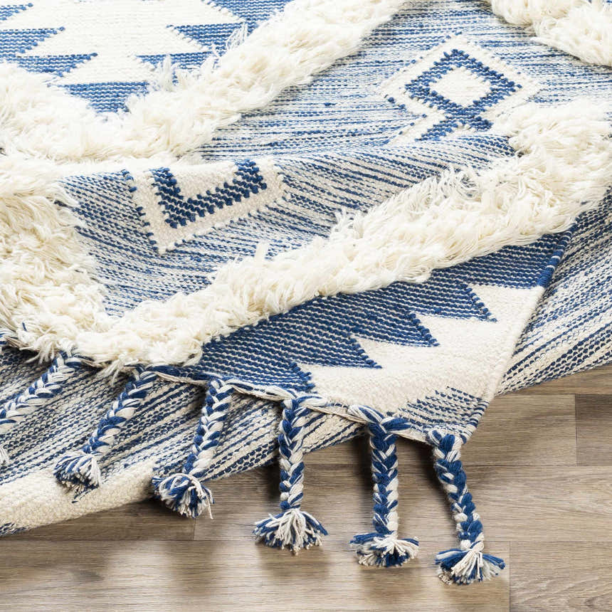 Close-up of the Painesdale Blue/White Wool Carpet. Hand-woven rug with geometric pattern, fringe, and blue/white braided tassels. Fold