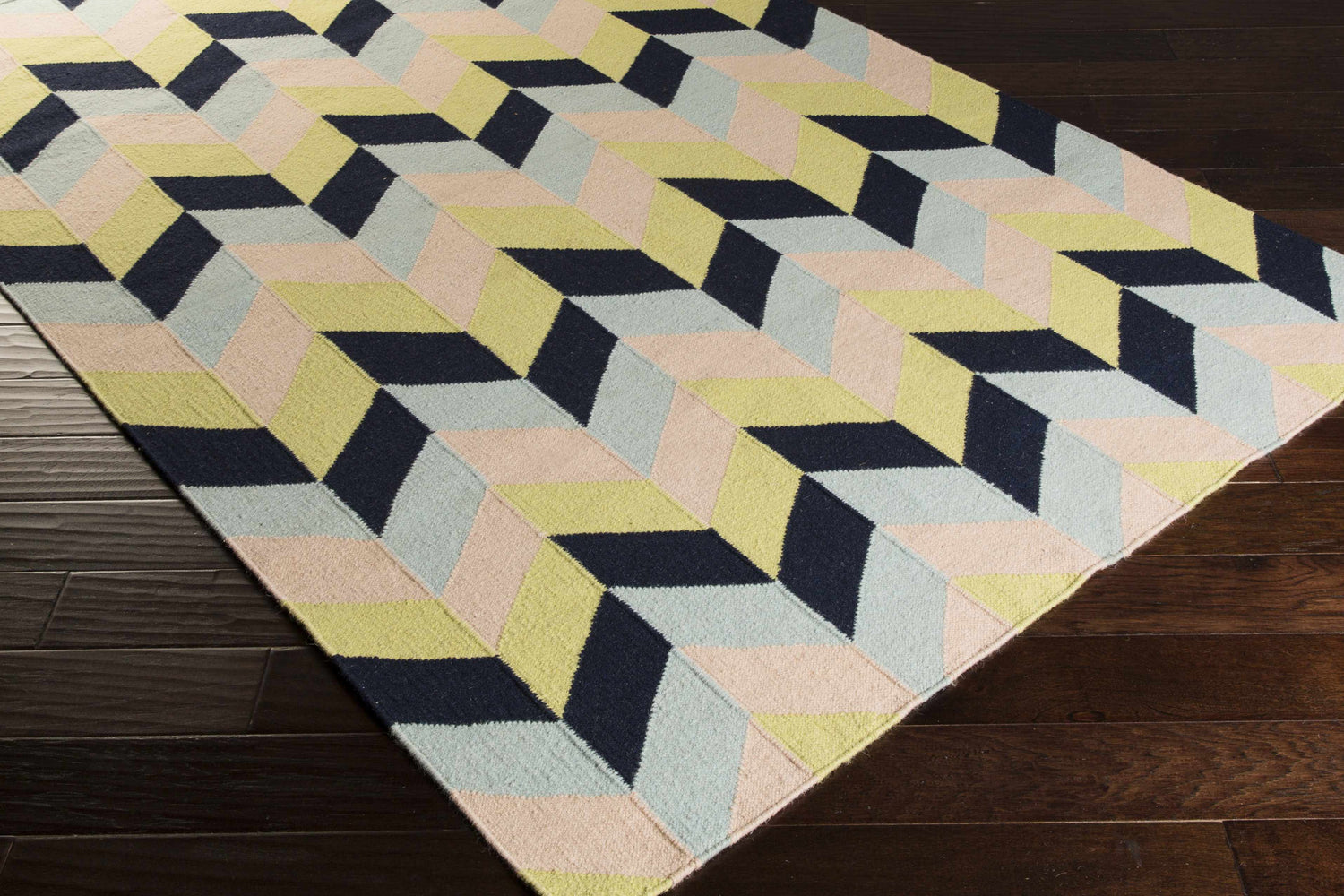 Close-up of the hand woven Pojoaque Area Carpet. Features a geometric design in dark blue, dusty pink, and light olive hues. Corner