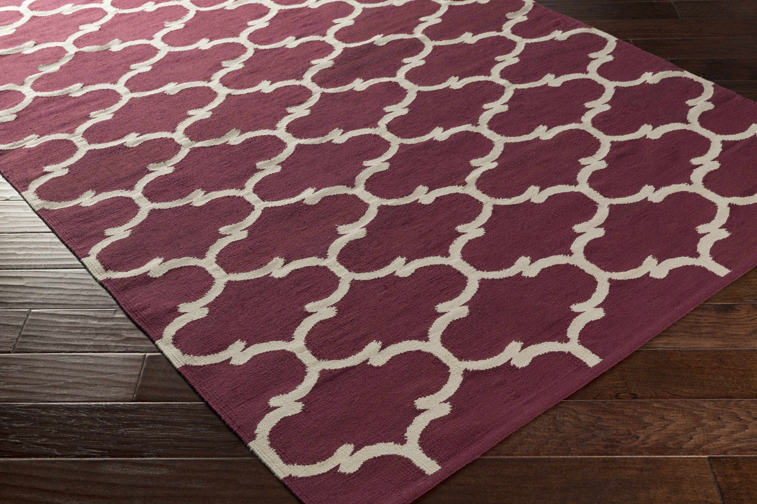 Close-up of the Racine Area Carpet. Dark purple cotton rug with an ivory quatrefoil pattern on a dark wood floor. Corner