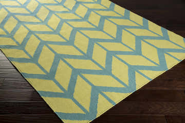 Close-up of the Readington Area Carpet. Hand-woven wool rug in olive green and teal geometric pattern on dark wood floor. Corner