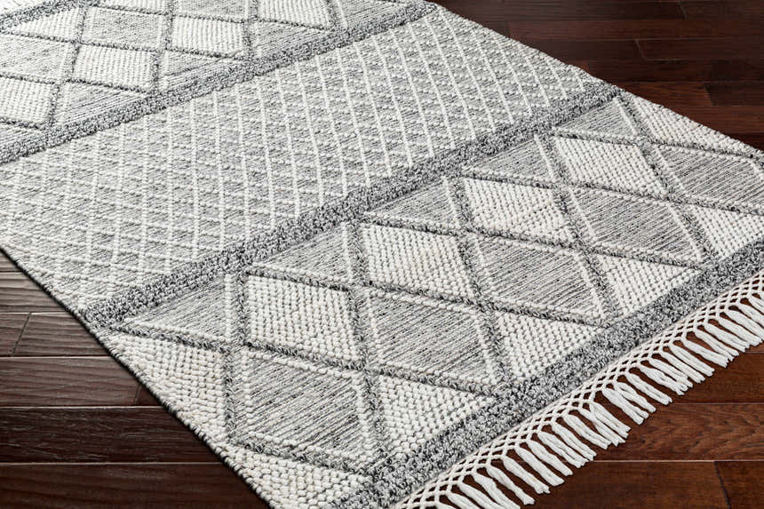 Close-up of the Rehn Area Rug: dark gray and light gray diamond pattern, hand woven with fringe detail on dark wood floor. Corner