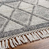 Close-up of the Rehn Area Rug, a hand-woven rug with a geometric diamond pattern and fringe, in dark gray, light gray, and taupe. Texture