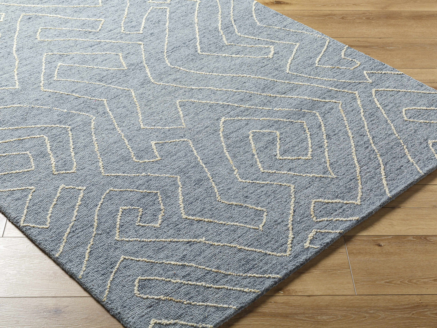 Close-up of Raffi Slate Blue Area Rug showing geometric design and textured wool on hardwood floor. Corner