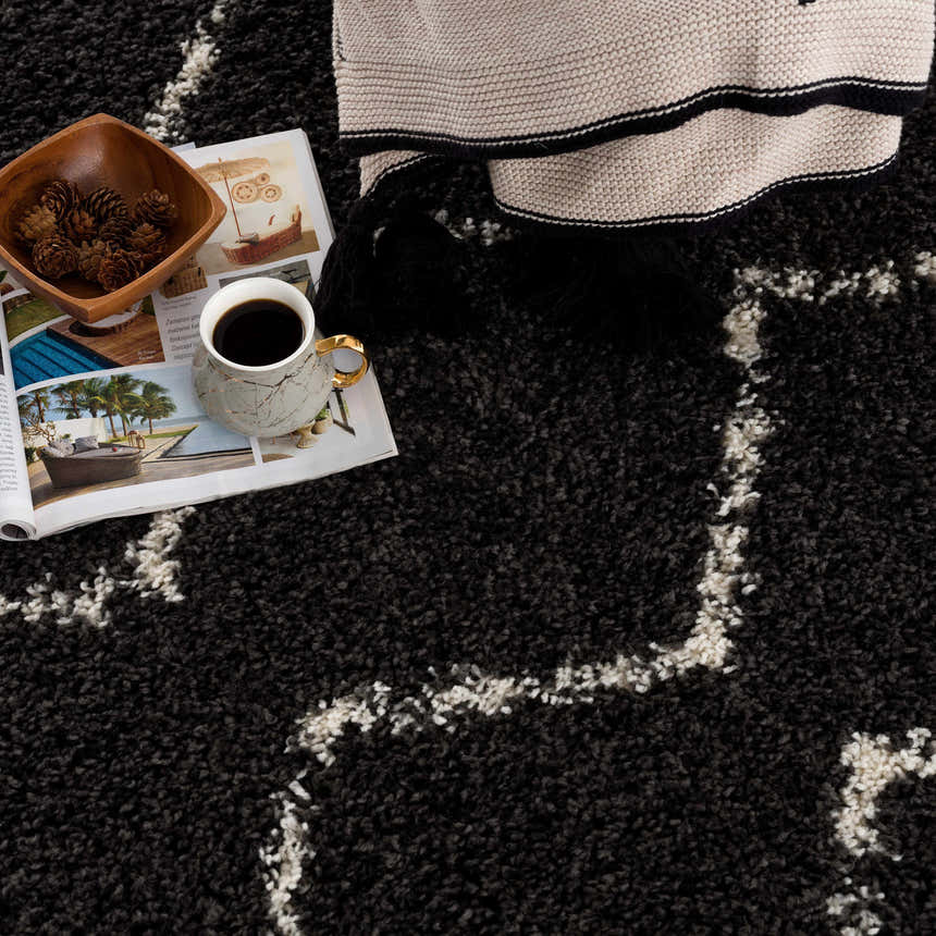 Close-up of the charcoal Ennis Area Rug with a light beige geometric pattern and shag texture. Styled with a coffee cup and blanket. Styleshot