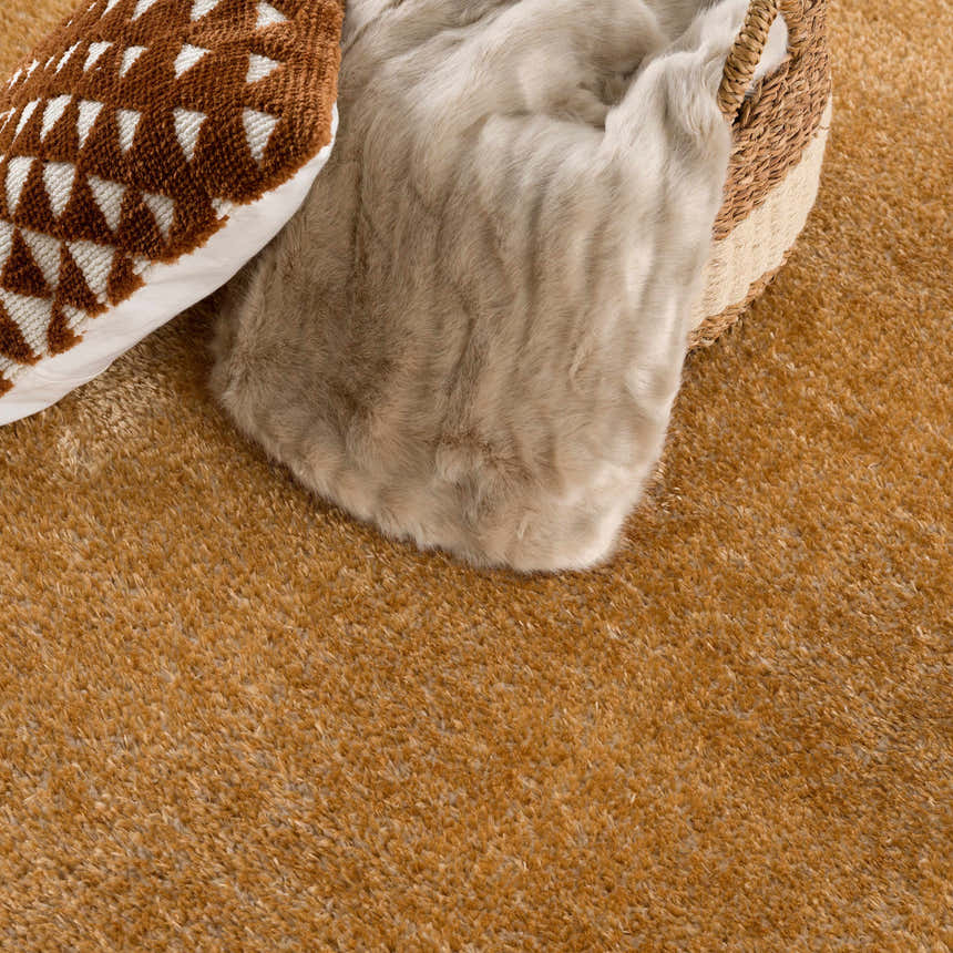 Close-up of the Heavenly Solid Mustard Plush Rug showing texture, with a throw blanket, patterned pillow, and woven basket. Styleshot