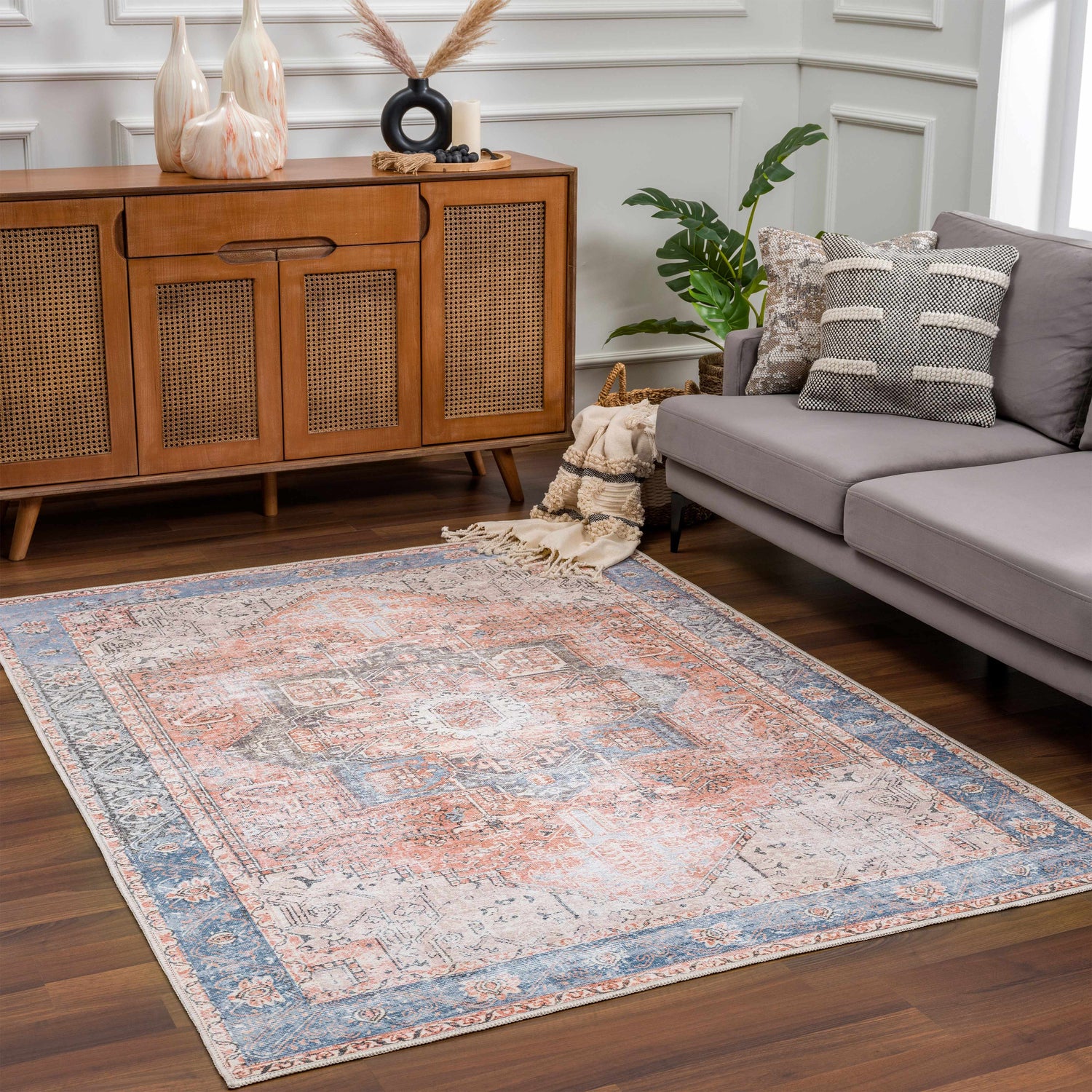 Living room scene with a Rosman Distressed Washable Area Rug in beige, blue, and brick red, styled with a gray sofa and wood cabinet. Roomscene-Rectangle