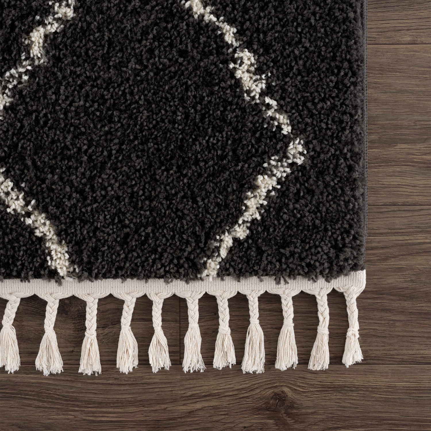 Close-up of the Ennis Area Rug, a charcoal shag rug with light beige diamond pattern and tassels on a wood floor. Swatch