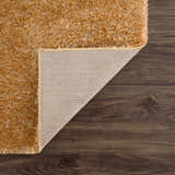 Close-up of Heavenly Solid Mustard Plush Rug showing texture, backing, and edge on a dark wood floor. Fold