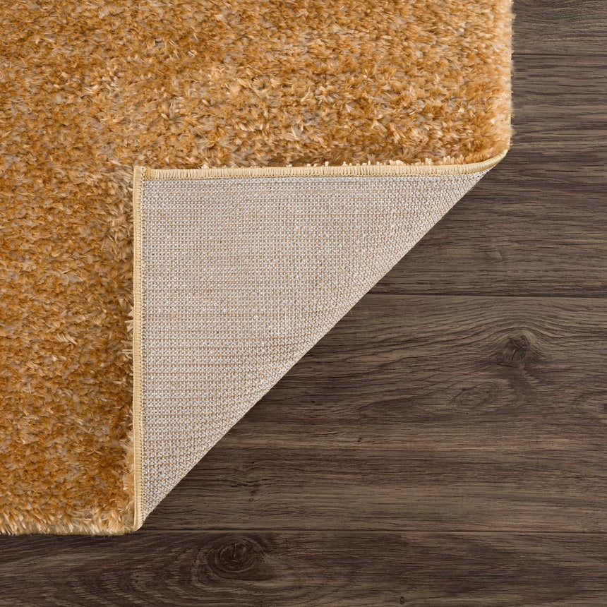 Close-up of Heavenly Solid Mustard Plush Rug showing texture, backing, and edge on a dark wood floor. Fold