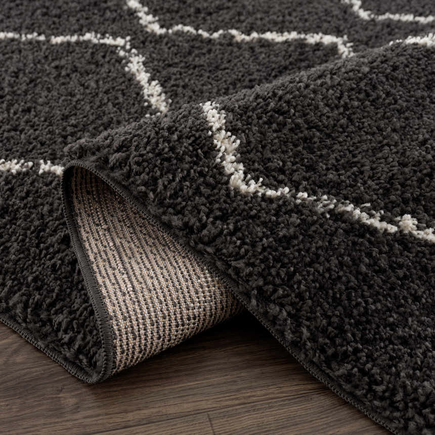 Close-up of the Ennis Area Rug. Charcoal shag rug with light beige geometric pattern and woven backing detail. Fold