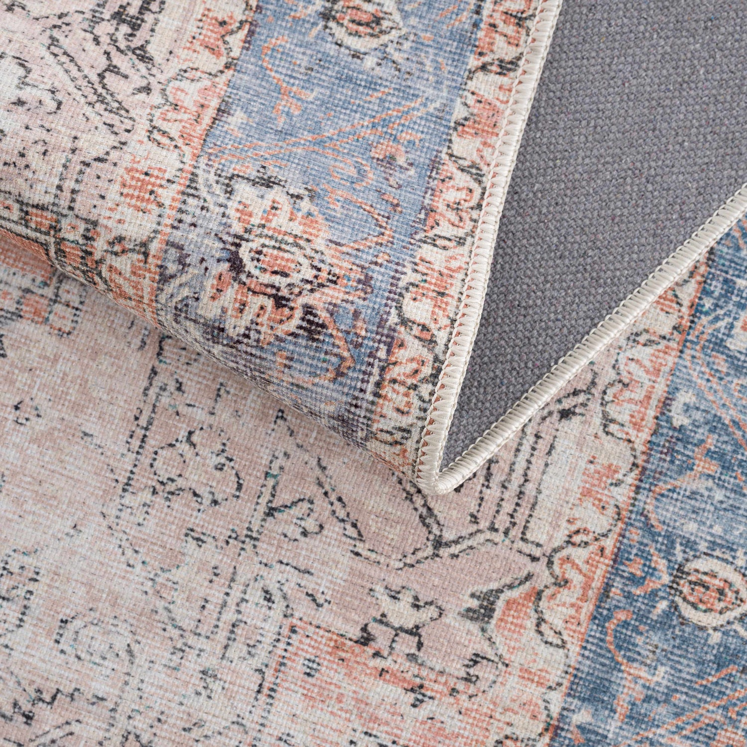 Close-up of the Rosman Distressed Washable Area Rug showing its beige, blue, and brick red oriental pattern and gray backing. Fold