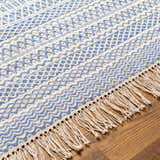 Close-up of the Sanc Blue/Cream rug showing its flatweave texture, geometric pattern, and fringe detail on a wood floor. Texture