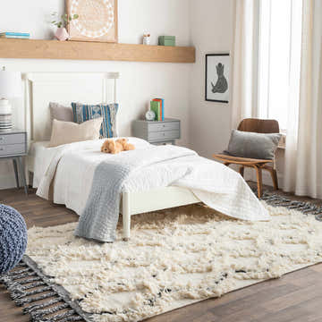 Bedroom scene with a white bed and the cream and black Sangerville Area Rug, a hand woven wool rug with tassels. Roomscene-Rectangle