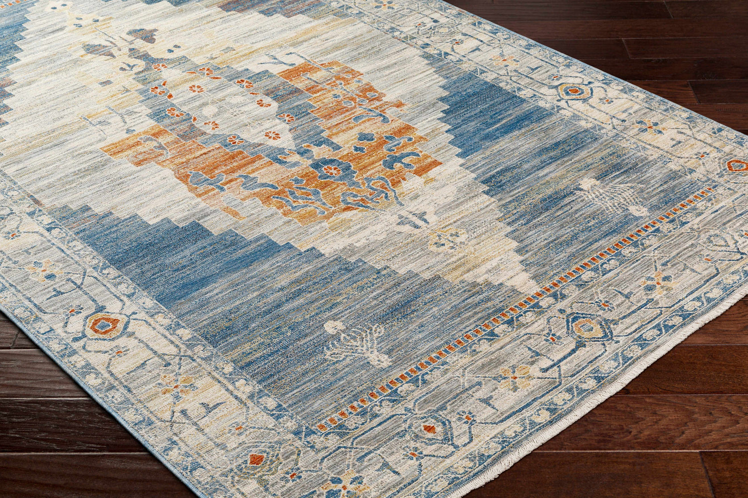 Close-up of the Shin Area Rug's traditional farmhouse design with clay, copper, denim, and gray hues on a dark wood floor. Corner
