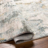 Close-up of the textured Adlai Rug with Fringes, showing its abstract pattern in dusty sage, ink blue, and off-white. Fold