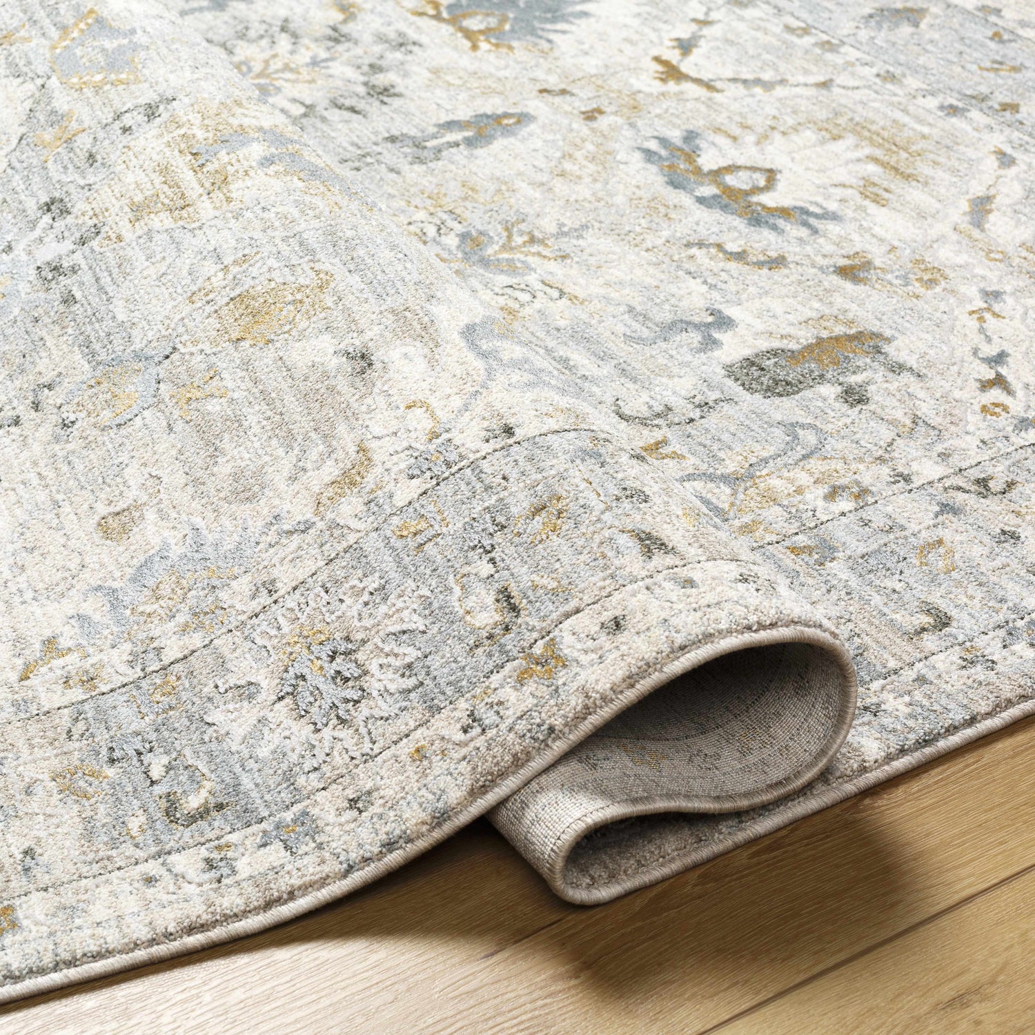 Close-up of the Emile Textured Rug, showing its light gray, taupe, and mustard traditional pattern and soft, textured weave. Fold