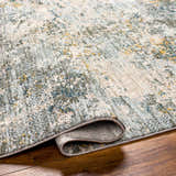 Close-up of the Pizi Textured Rug with Fringes in dusty sage, ink blue, and mustard on a wood floor. Contemporary style. Fold