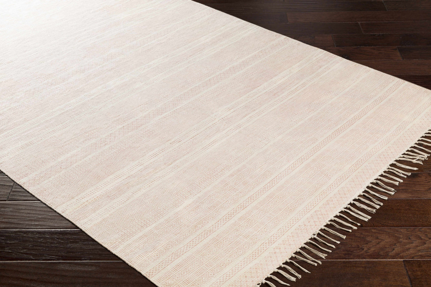 Close-up of Serena Area Rug in dusty coral and peach cotton with woven stripes and fringe on dark wood floor. Corner