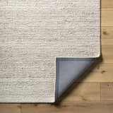 Close-up of the hand-tufted Urmat Wool Area Rug in light silver and warm gray on a wood floor, showing rug backing. Fold