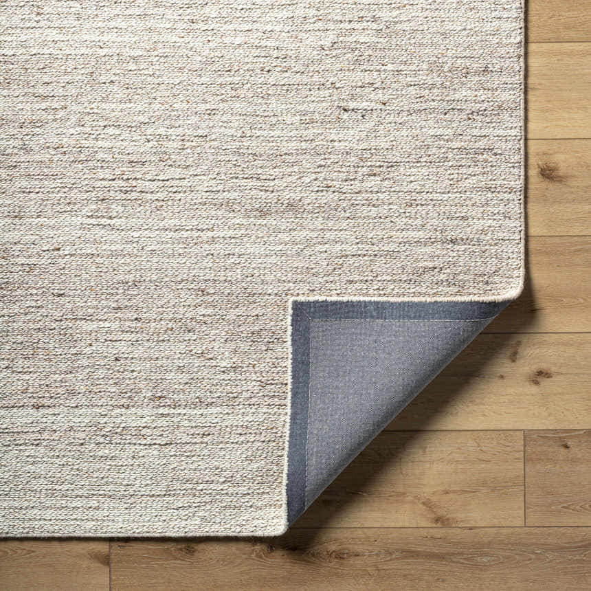 Close-up of the hand-tufted Urmat Wool Area Rug in light silver and warm gray on a wood floor, showing rug backing. Fold