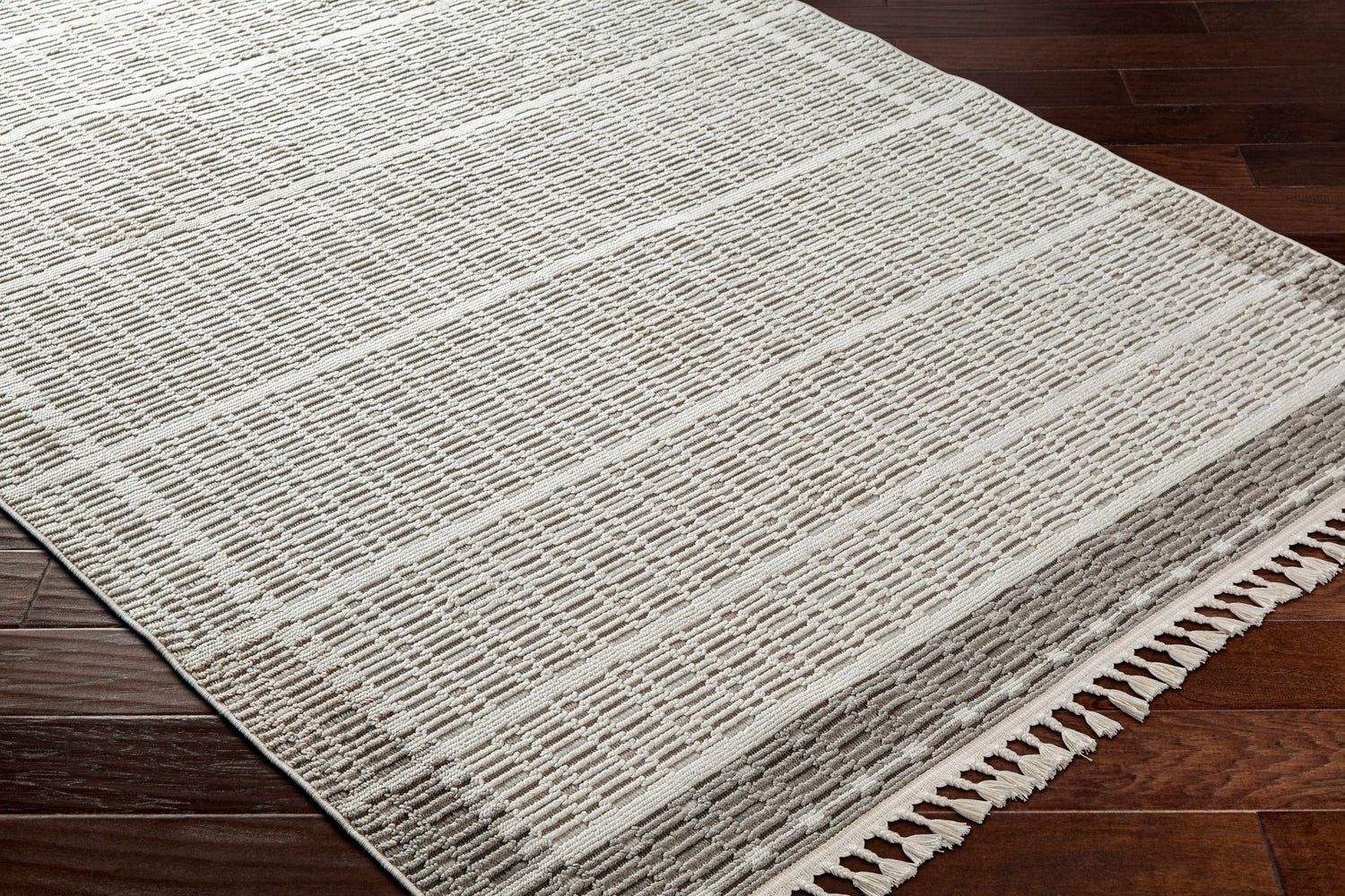 Close-up of the beige and gray Ajda Area Rug showing its geometric pattern and tassel fringe on dark wood flooring. Corner