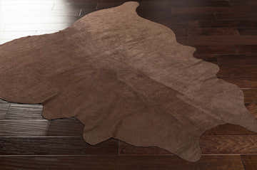 Close-up of the brown Sitka 5x7 Cowhide Rug on a dark brown hardwood floor. The leather rug has a soft, suede-like texture. Corner