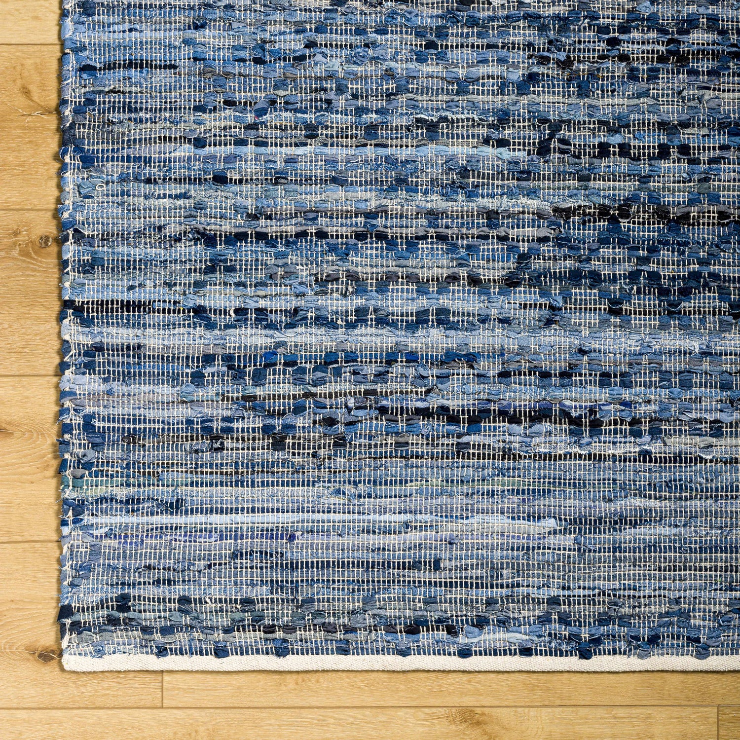 Close-up of the hand woven Goro Area Rug. Features charcoal, gray, and silver tones on a light wood floor. Corner